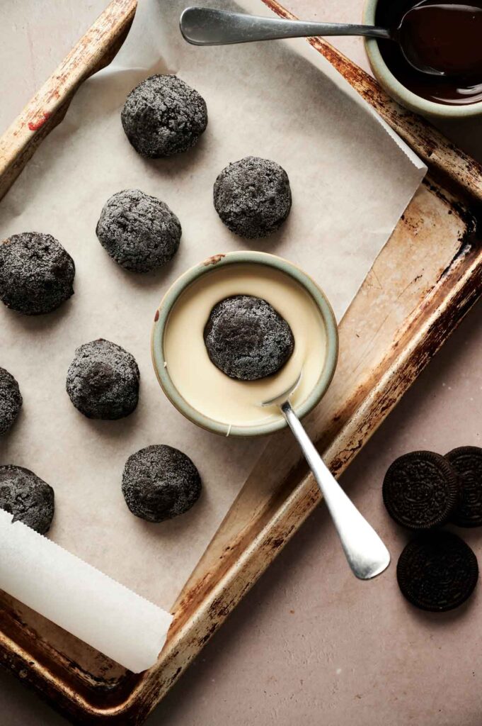 A baking tray with Oreo Balls and chocolate cookie dough balls on parchment paper; one treat is being dipped in white chocolate in a small bowl with a spoon.