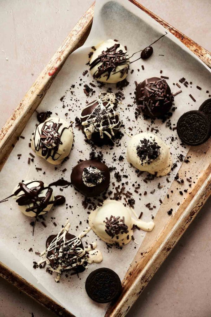 A baking tray with nine Oreo Balls—truffles coated in white and dark chocolate, drizzled with chocolate and sprinkled with cookie crumbs—sits on parchment paper. Two Oreo cookies are on the tray.