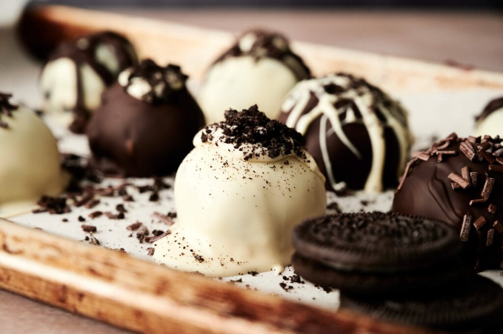 A close-up of assorted chocolate truffles, some resembling Oreo Balls coated in white and dark chocolate, garnished with cookie crumbs and sprinkles, arranged on a tray.
