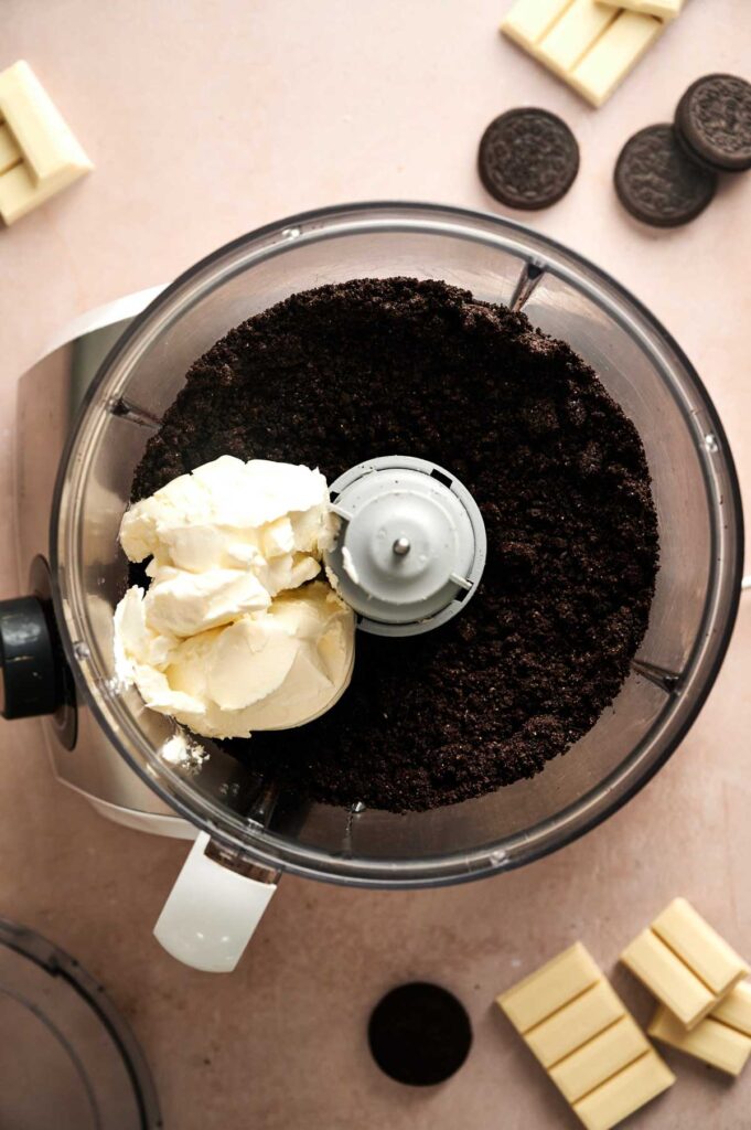 A food processor with crushed chocolate cookies and cream cheese inside, ready to be transformed into delicious Oreo Balls, is surrounded by whole cookies and white chocolate pieces on a light countertop.
