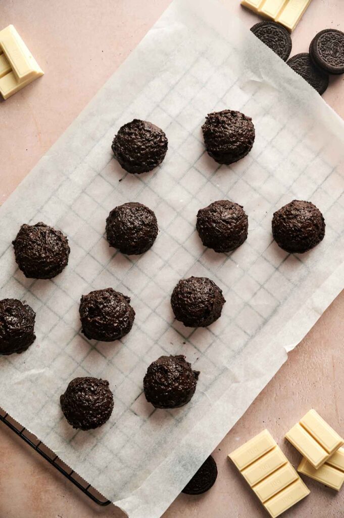 Ten scoops of chocolate cookie dough on a parchment-lined cooling rack, with white chocolate pieces, sandwich cookies, and homemade Oreo Balls nearby.
