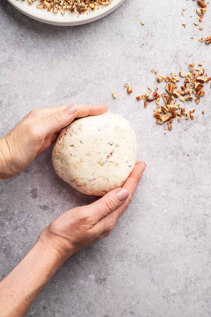 Two hands holding a round pineapple cheese ball with herbs on a gray surface, next to a small pile of chopped nuts.