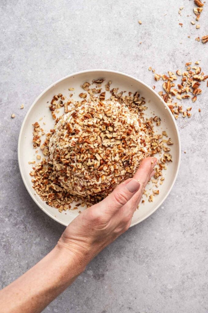 A hand holds a round Pineapple Cheese Ball covered in chopped pecans on a white plate, with scattered pecans around it.