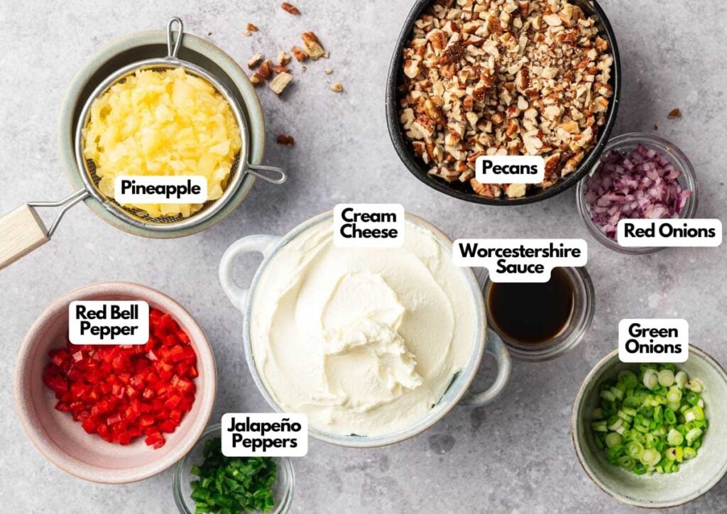 A top-down view of bowls holding ingredients for Pineapple Cheese Ball: chopped pineapple, pecans, red onions, red bell pepper, green onions, jalape&ntilde;o peppers, cream cheese, and Worcestershire sauce&mdash;each bowl is clearly labeled.