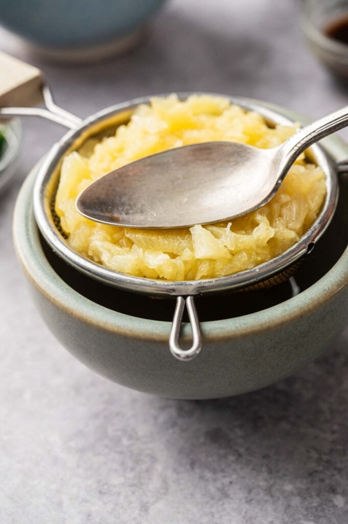 Crushed pineapple being pressed with a spoon in a small mesh strainer set over a bowl to drain excess juice.