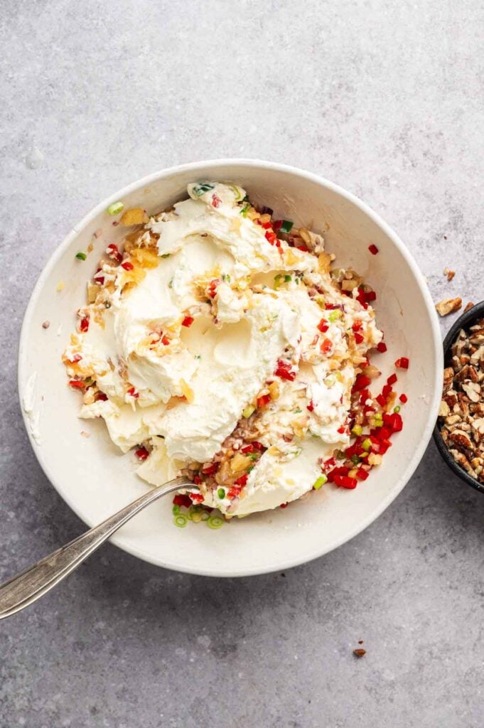 A bowl of cream cheese mixed with chopped red peppers, green onions, and pecans—the classic base for a pineapple cheese ball—sits ready with a spoon in the bowl and extra pecans on the side.