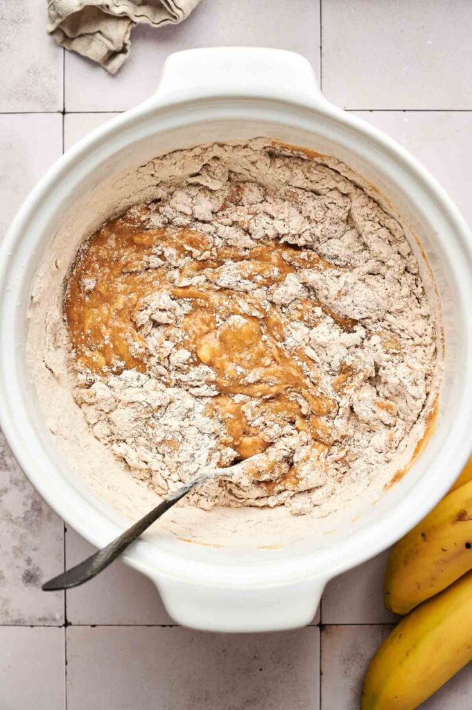 A white mixing bowl with flour and wet ingredients being combined with a spoon, next to two bananas on a tiled countertop—perfect for preparing Pumpkin Banana Bread.