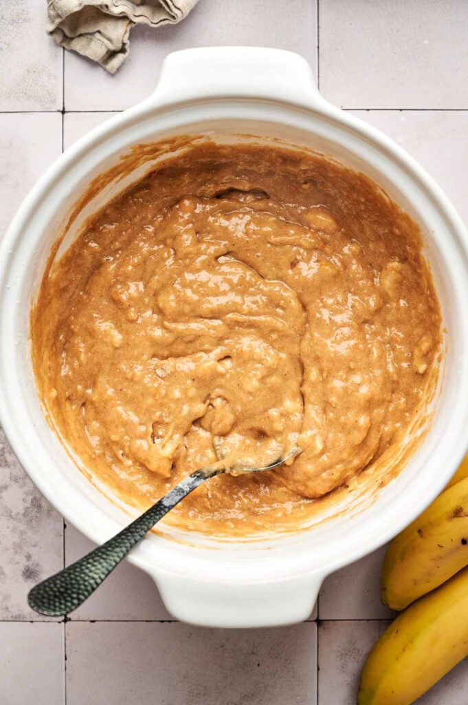 A white bowl filled with Pumpkin Banana Bread batter and a spoon, next to two whole bananas on a tiled surface.