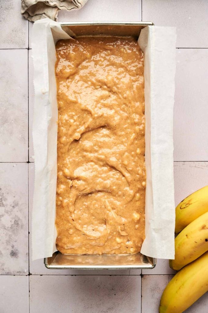 A loaf pan lined with parchment paper filled with Pumpkin Banana Bread batter, with two bananas placed beside the pan on a tiled surface.