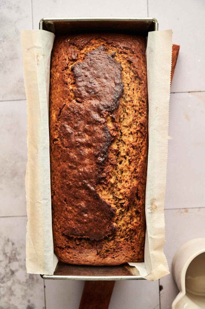 A loaf of Pumpkin Banana Bread in a parchment-lined rectangular metal pan sits on a tiled surface.
