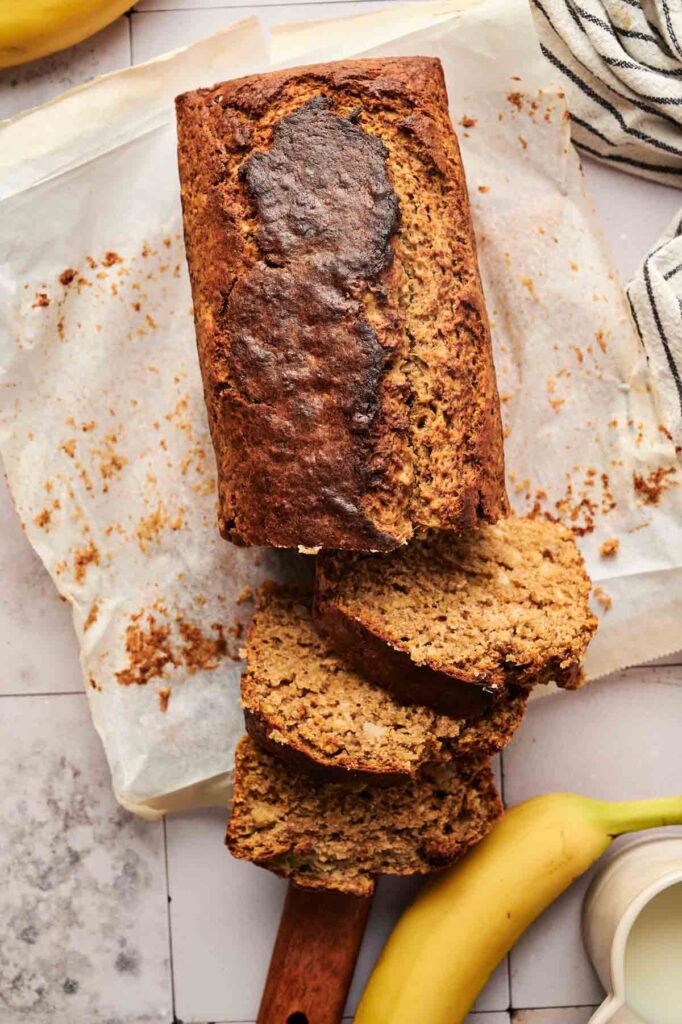 A loaf of Pumpkin Banana Bread on parchment paper, partially sliced, with a banana and a striped towel beside it.