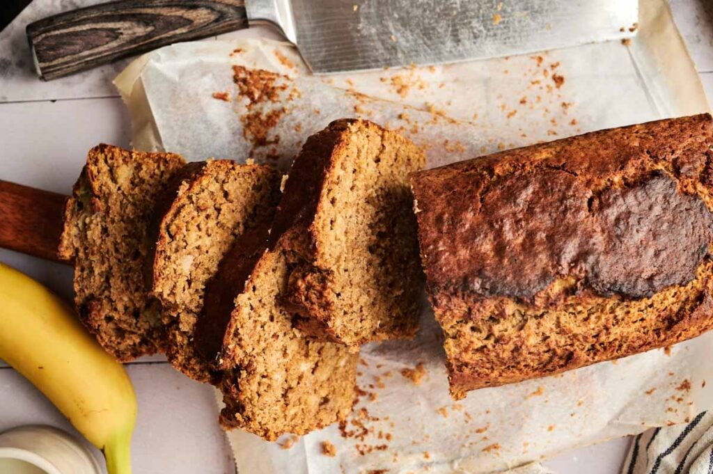 A loaf of Pumpkin Banana Bread, partially sliced, sits on parchment paper next to a banana and a knife.
