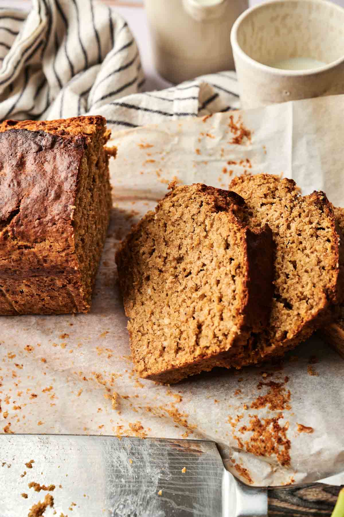 A partially sliced loaf of Pumpkin Banana Bread on parchment paper, with two slices cut and a knife nearby; a striped cloth and a cup are in the background.