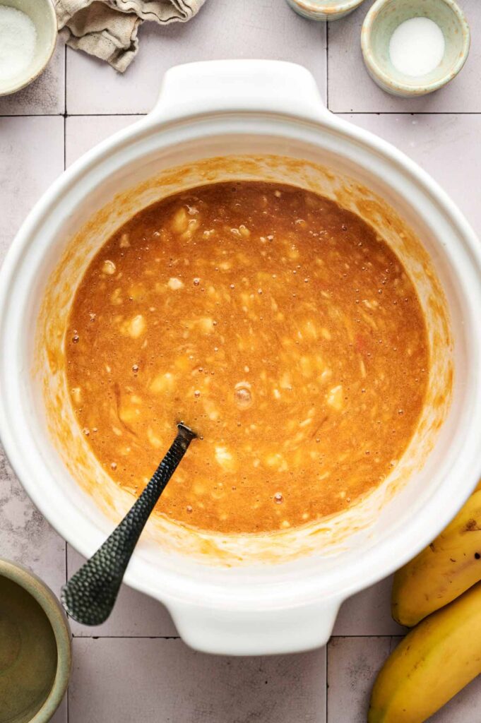 A white mixing bowl filled with Pumpkin Banana Bread batter and a spoon, surrounded by ingredients and bananas on a tiled surface.