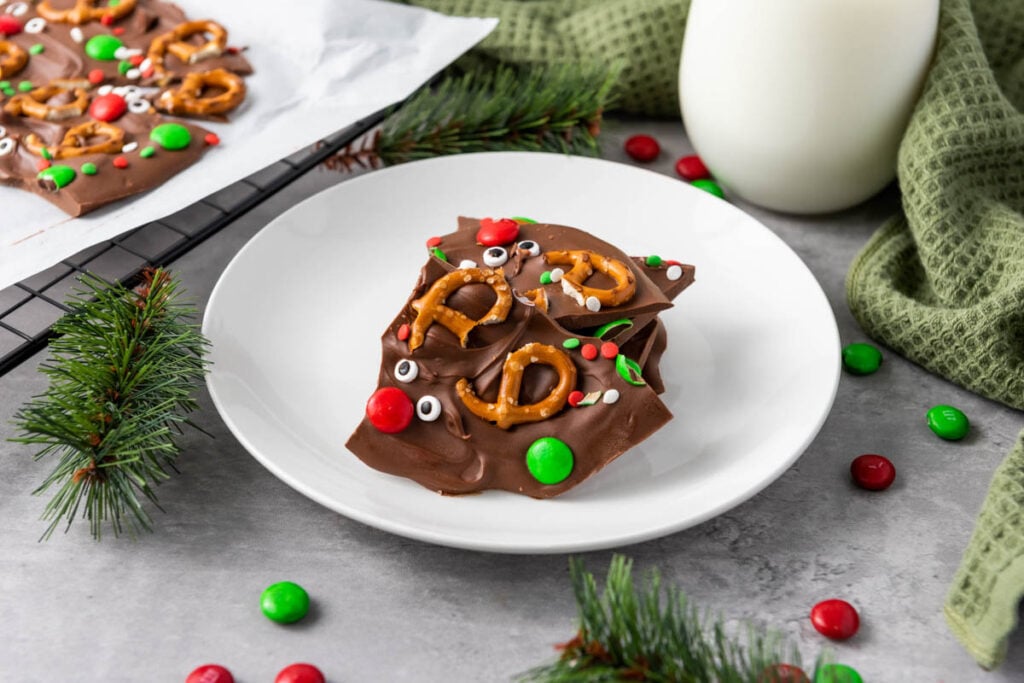 Reindeer Chocolate Bark topped with pretzels, candy eyes, and festive red and green candies sits on a white plate, surrounded by holiday decorations and a glass of milk in the background.