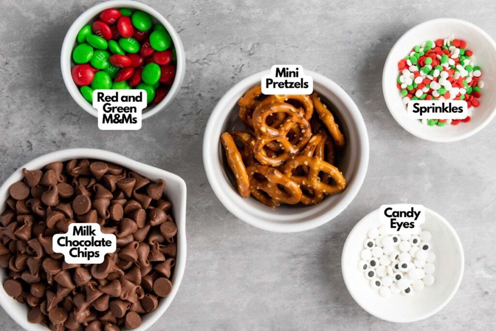 Five bowls with ingredients for Reindeer Chocolate Bark—red and green M&Ms, mini pretzels, sprinkles, milk chocolate chips, and candy eyes—are arranged on a gray surface.