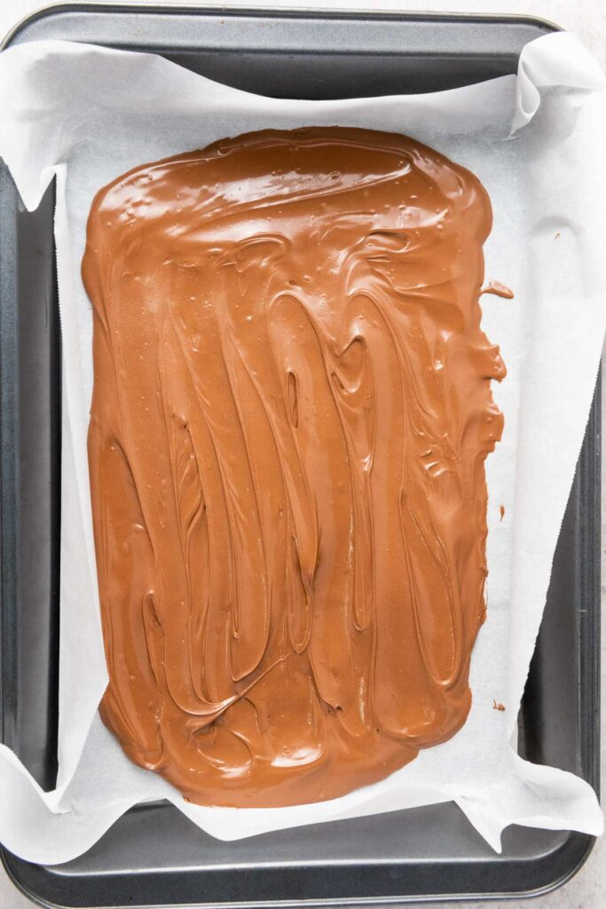 A baking pan lined with parchment paper holds a layer of melted milk chocolate spread evenly across the surface, ready to be transformed into festive Reindeer Chocolate Bark.