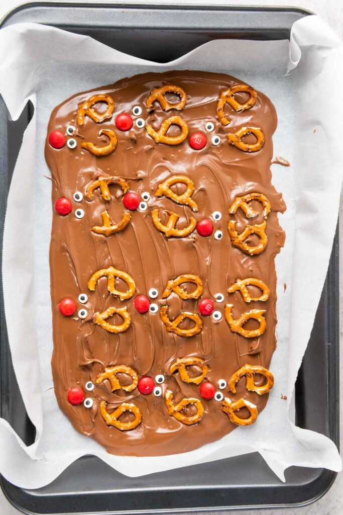 Rectangular tray of Reindeer Chocolate Bark topped with pretzels, red candies, and candy eyes, arranged to resemble reindeer faces, on parchment paper.