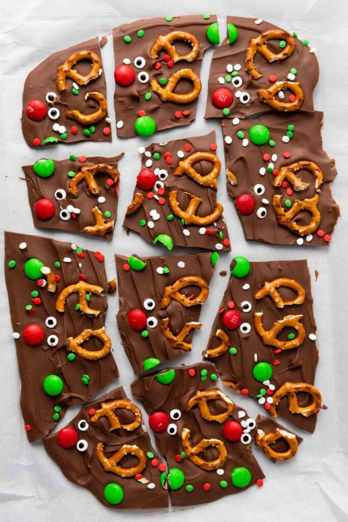 Reindeer Chocolate Bark broken into pieces, decorated with pretzels, red and green candy-coated chocolates, candy eyes, and holiday-themed sprinkles on a parchment paper background.