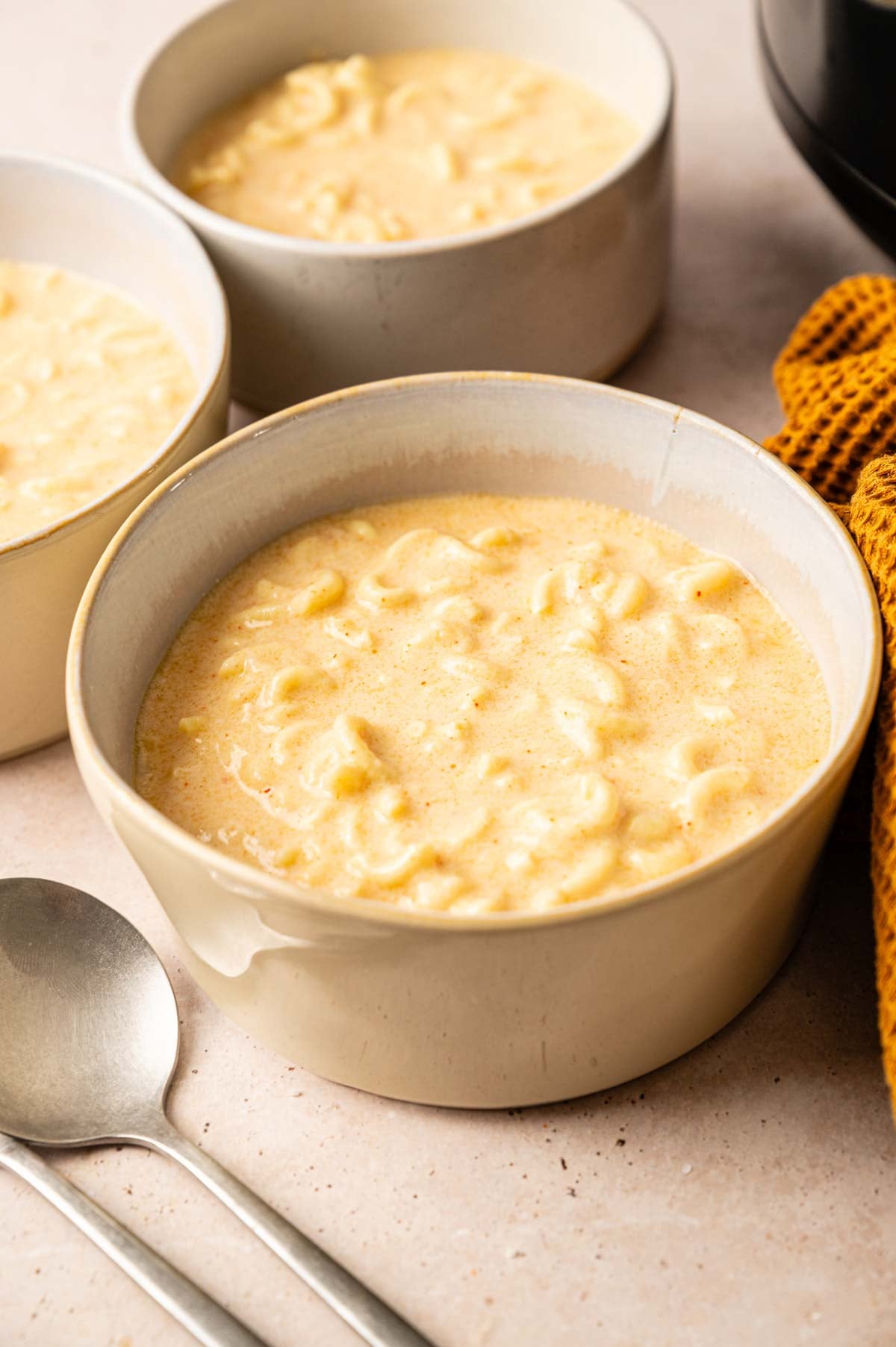 Three bowls of creamy Slow Cooker Mac and Cheese are placed on a light surface next to two metal spoons and a textured yellow cloth.