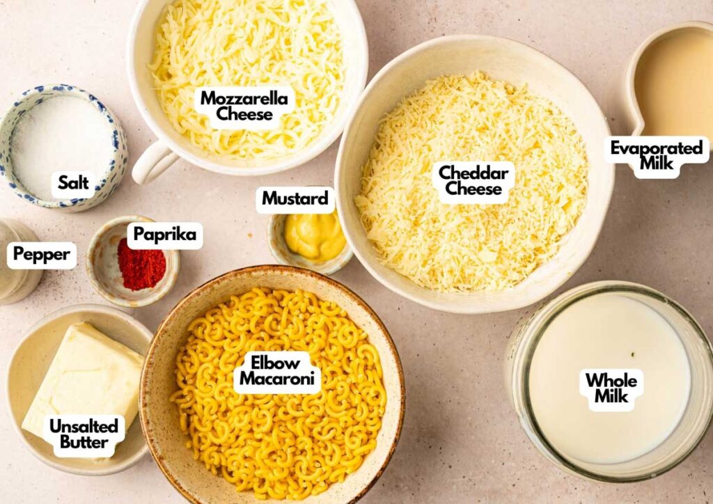 Bowls containing ingredients for Slow Cooker Mac and Cheese&mdash;shredded mozzarella and cheddar cheese, evaporated milk, whole milk, elbow macaroni, unsalted butter, salt, pepper, paprika, and mustard&mdash;are arranged on a countertop.