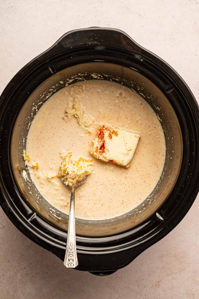 A slow cooker filled with Slow Cooker Mac and Cheese&mdash;creamy mixture, shredded cheese, a block of cream cheese, and a sprinkle of paprika. A metal spoon rests inside the mixture.