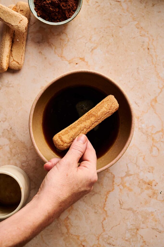 A hand dips a ladyfinger biscuit into a bowl of dark liquid, with tiramisu brownies, more ladyfingers, a jug, and a bowl of cocoa powder nearby on a marble countertop.