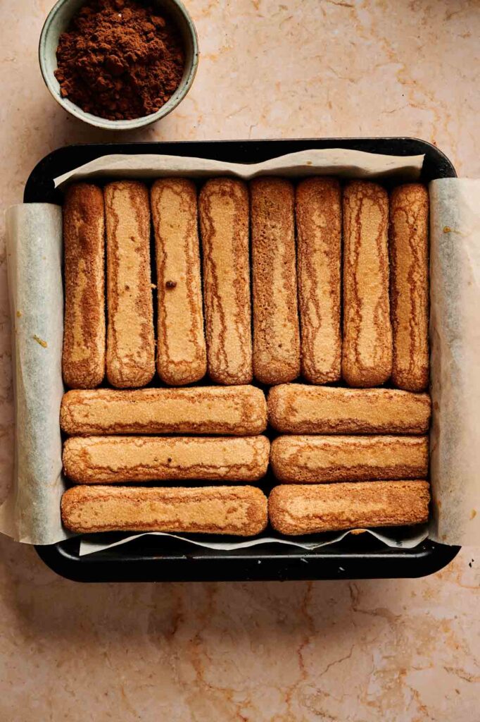 Ladyfinger cookies are arranged over a brownie layer in a parchment-lined baking pan, layered lengthwise and crosswise, with a bowl of cocoa powder nearby.
