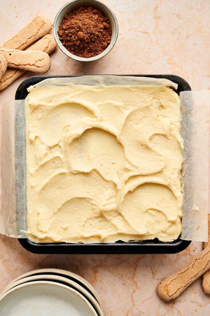 A square baking pan lined with parchment paper and filled with smooth, swirled tiramisu brownies sits on a countertop, surrounded by cocoa powder, ladyfingers, and stacked plates.