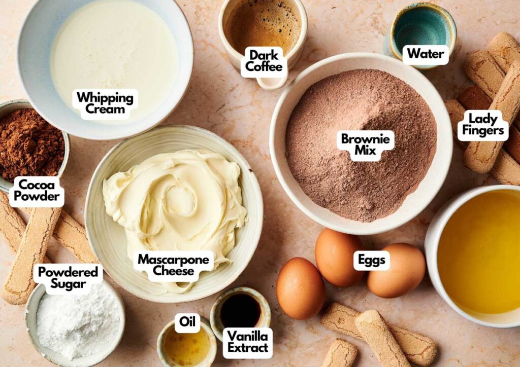 Various dessert ingredients for Tiramisu Brownies are arranged on a countertop, each labeled: whipping cream, dark coffee, water, lady fingers, brownie mix, eggs, oil, vanilla extract, mascarpone cheese, powdered sugar, and cocoa powder.