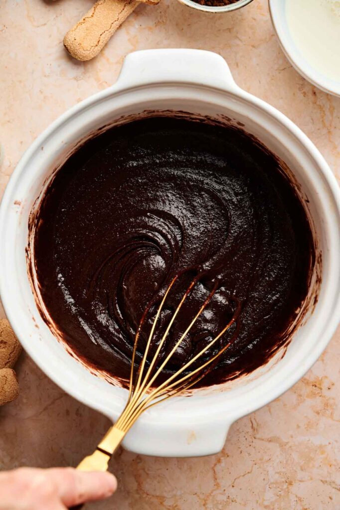 A hand holds a gold whisk mixing thick chocolate batter for tiramisu brownies in a white bowl on a marble countertop, with cookie pieces and a bowl nearby.