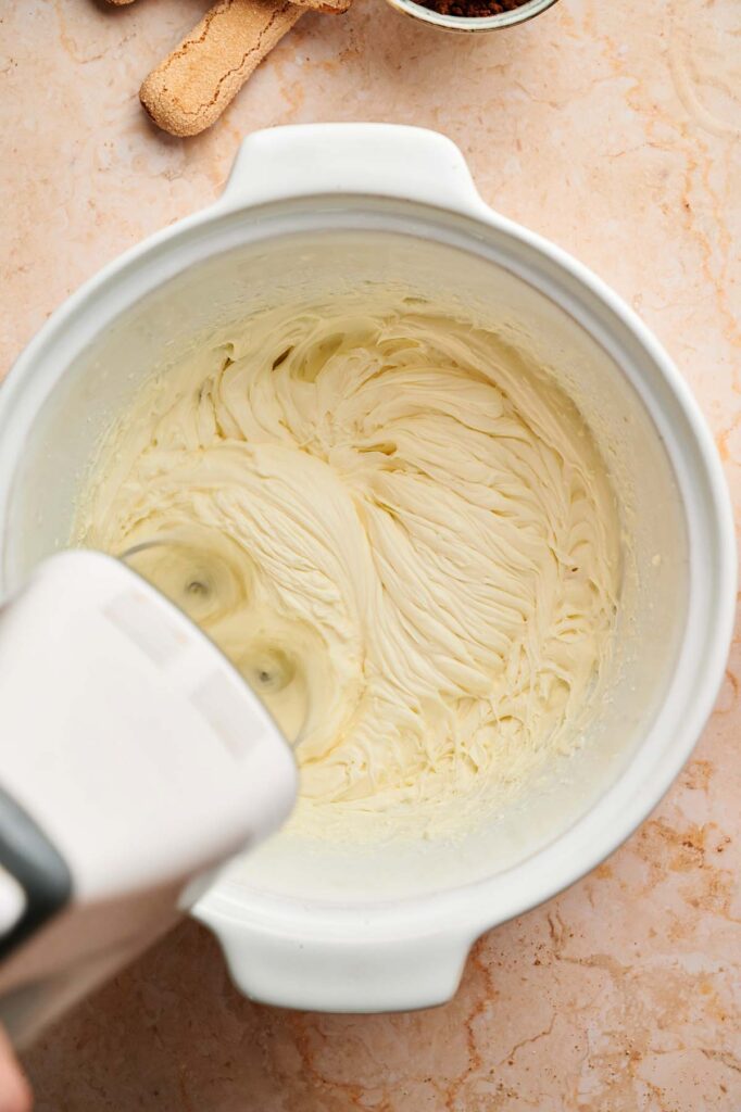 Electric hand mixer blending creamy batter in a white bowl on a beige countertop, with a few ladyfinger biscuits nearby—perfect for whipping up decadent Tiramisu Brownies.