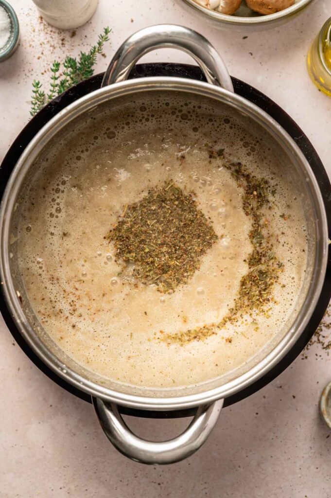 A pot of simmering soup with seasonings and herbs sprinkled on top, surrounded by cooking ingredients on a light countertop&mdash;perfect for enjoying with a ladle of rich vegetarian gravy.