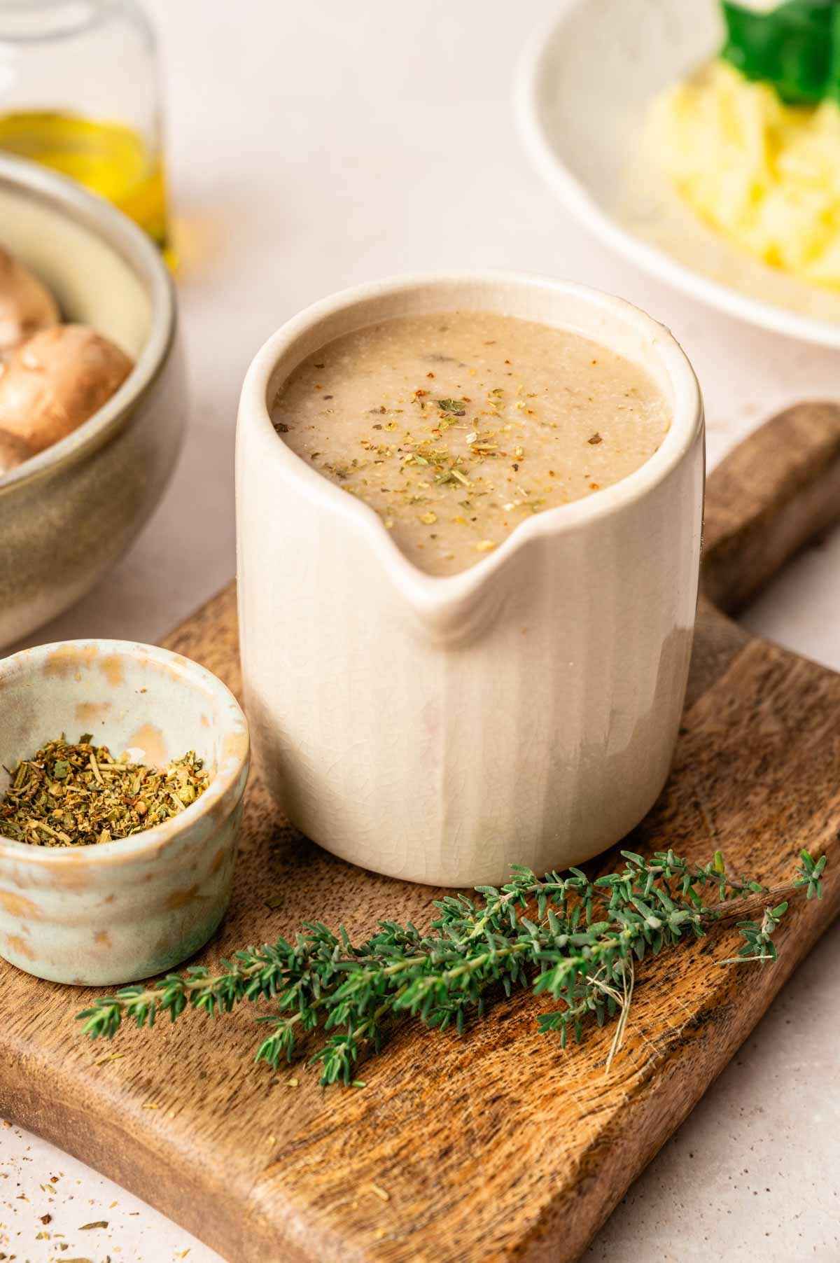 A ceramic gravy boat filled with creamy vegetarian mushroom gravy sits on a wooden board, next to a small bowl of dried herbs and fresh thyme.