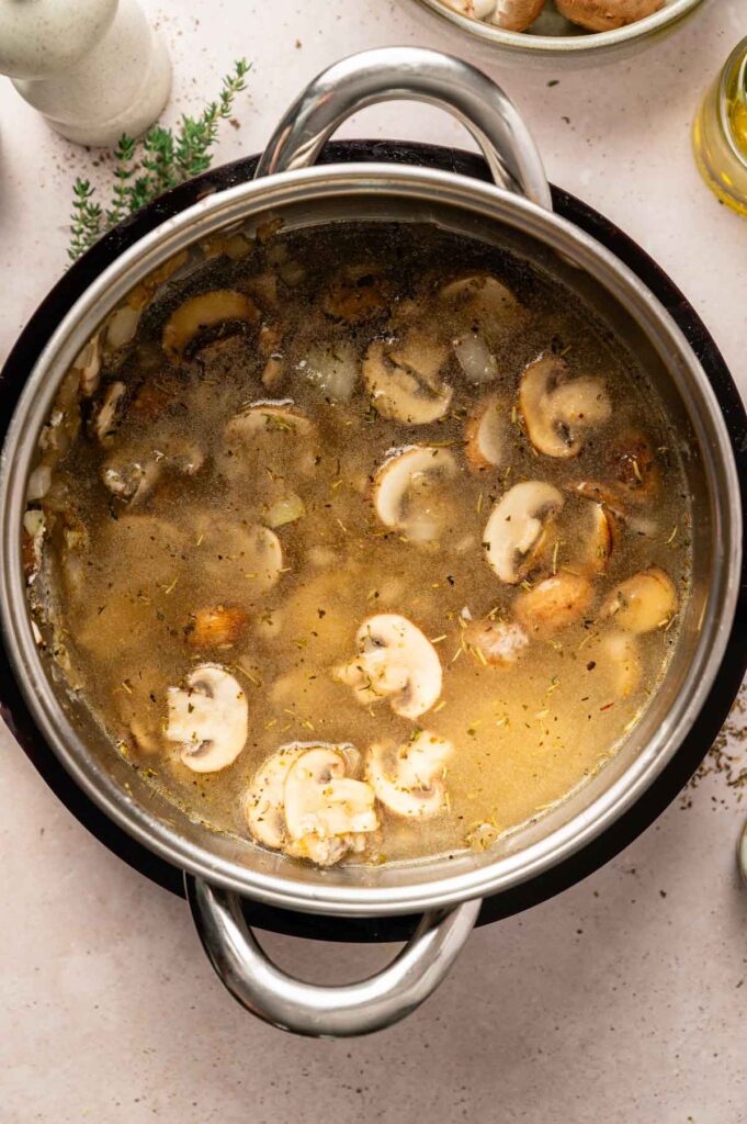 A pot of mushroom soup with sliced mushrooms and herbs simmering in broth, viewed from above on a light countertop—perfect for serving as a hearty vegetarian gravy.