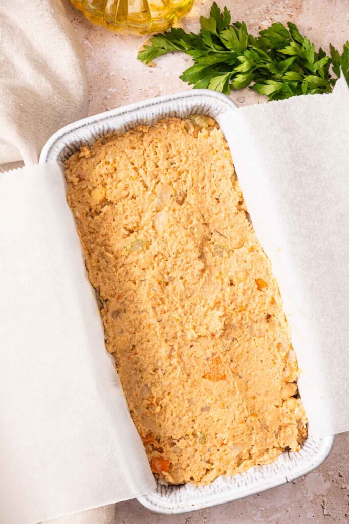 A rectangular baking dish lined with parchment paper holds an unbaked Vegetarian Meatloaf chickpea mixture, with fresh parsley and a bottle of oil in the background.