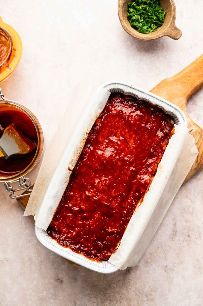 A Vegetarian Meatloaf covered in a reddish glaze sits in a parchment-lined loaf pan on a wooden board, with a jar of sauce and a small bowl of chopped herbs nearby.