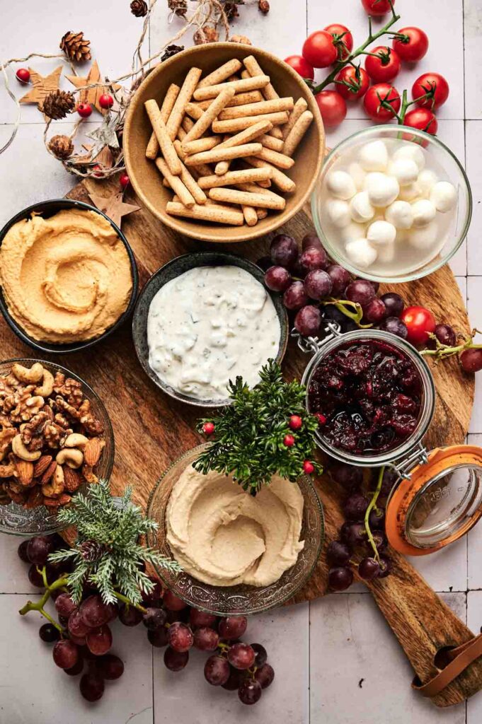 A Veggie Christmas Charcuterie Board features a wooden board with breadsticks, mozzarella balls, hummus, dip, walnuts, grape jelly, grapes, and cherry tomatoes artfully arranged around small bowls.