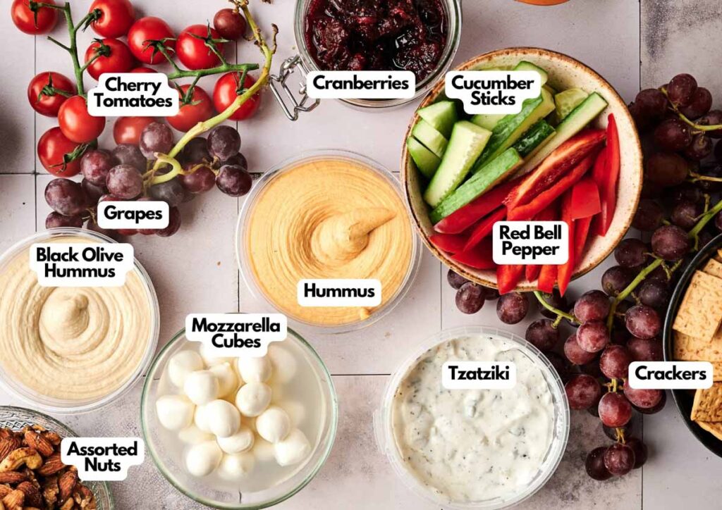 Enjoy a Veggie Christmas Charcuterie Board featuring cherry tomatoes, grapes, assorted nuts, hummus, black olive hummus, mozzarella cubes, tzatziki, cranberries, cucumber sticks, red bell pepper, and crackers.