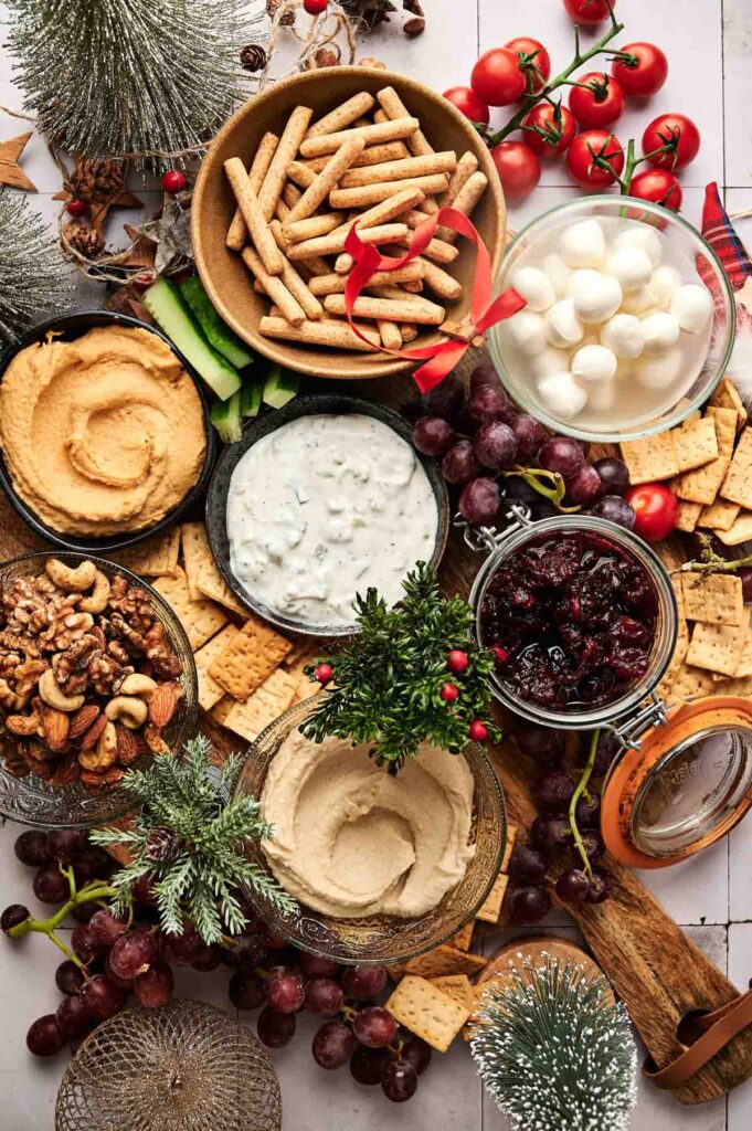 A festive Veggie Christmas Charcuterie Board features a colorful snack platter with dips, breadsticks, crackers, grapes, cherry tomatoes, mozzarella balls, mixed nuts, and a jar of cranberry sauce, all beautifully arranged and surrounded by cheerful holiday decorations.