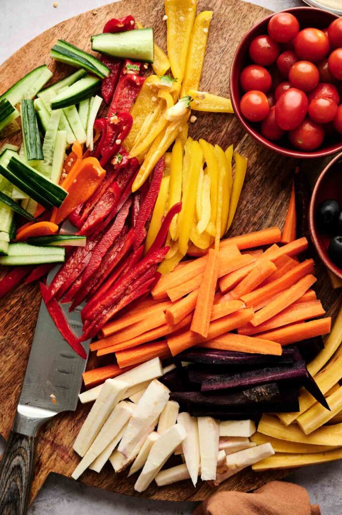 Assorted sliced vegetables including cucumbers, bell peppers, carrots, and parsnips are arranged on a wooden Veggie Thanksgiving Charcuterie Board, with bowls of cherry tomatoes and black olives nearby. A knife is also visible.