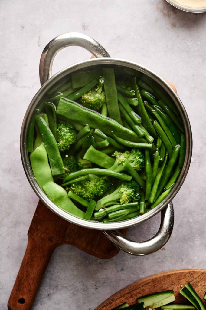A stainless steel pot filled with fresh green beans and broccoli submerged in water sits on a wooden cutting board, ready to be the perfect addition to your Veggie Thanksgiving Charcuterie Board.