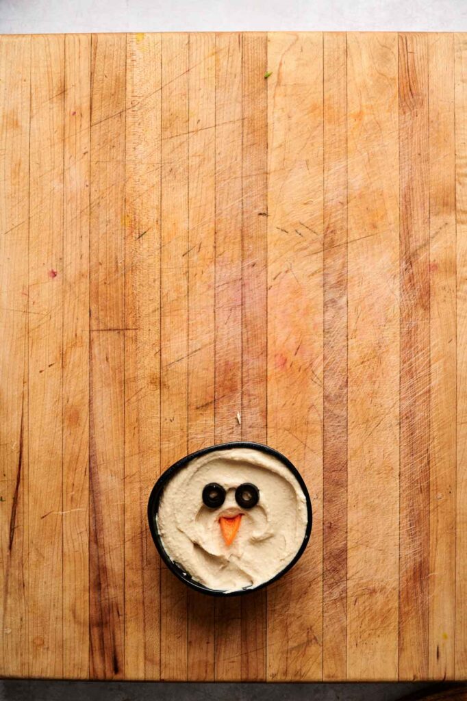 A small bowl of creamy dip on a wooden surface, decorated with two olive slices and a carrot triangle to resemble a face, makes a playful addition to any Veggie Thanksgiving Charcuterie Board.