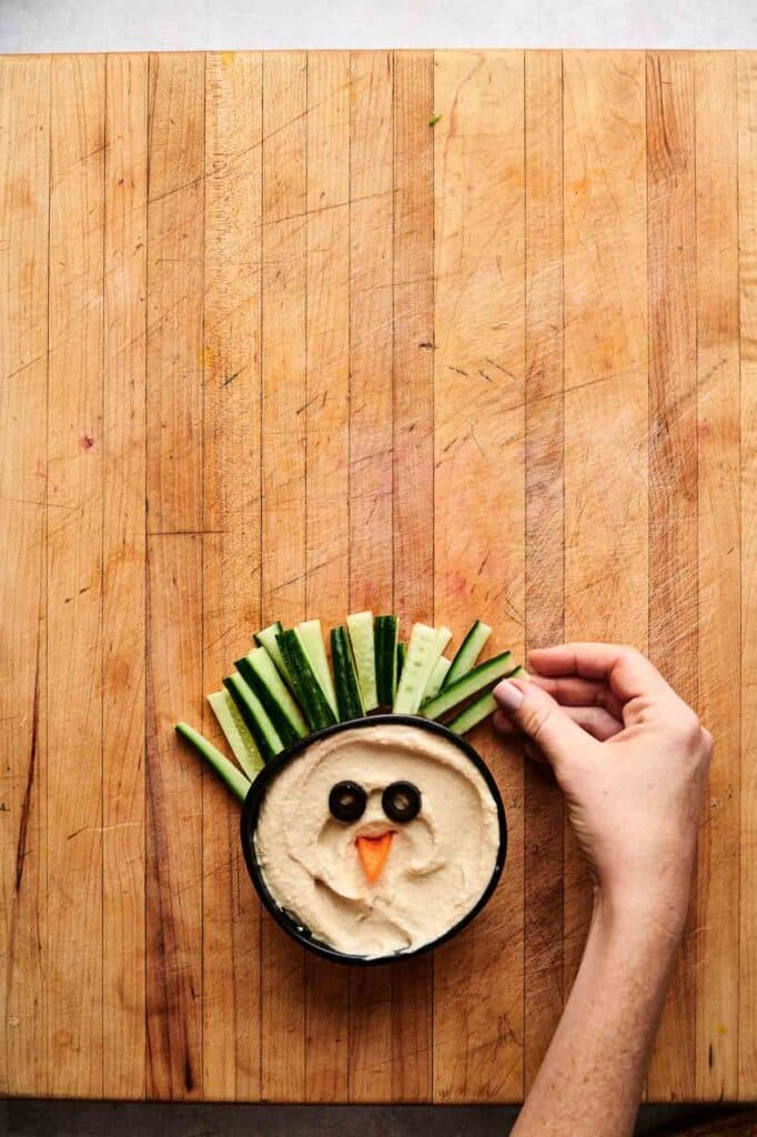 A hand places cucumber sticks in hummus to look like feathers. Perfect for a Veggie Thanksgiving Charcuterie Board, the dip is decorated with olive slices for eyes and a carrot slice for a beak, resembling a bird face.