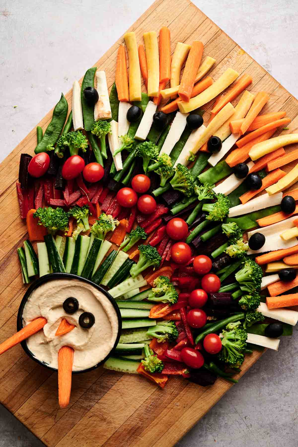 A Veggie Thanksgiving Charcuterie Board features a wooden board with assorted sliced vegetables arranged like a turkey, with a bowl of hummus as the head, complete with a carrot beak and olive eyes.