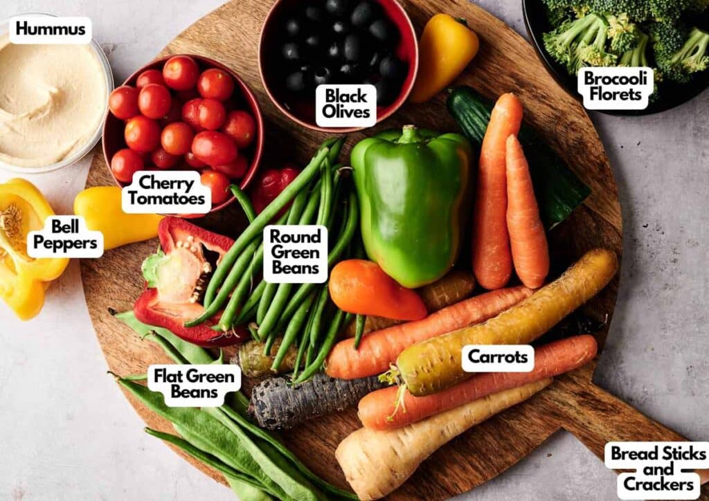 A Veggie Thanksgiving Charcuterie Board featuring hummus, cherry tomatoes, black olives, bell peppers, green beans, carrots, broccoli florets, and assorted bread sticks and crackers&mdash;each item beautifully labeled.