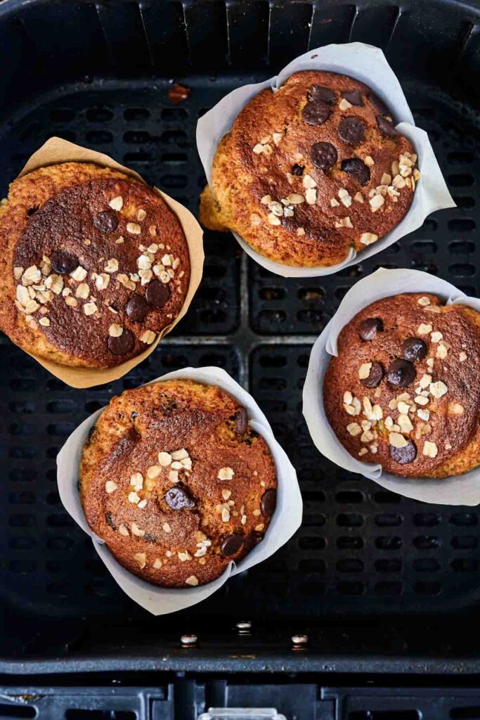 Four Air Fryer Banana Chocolate Chip Muffins with oats on top, nestled in an air fryer basket lined with white parchment paper.