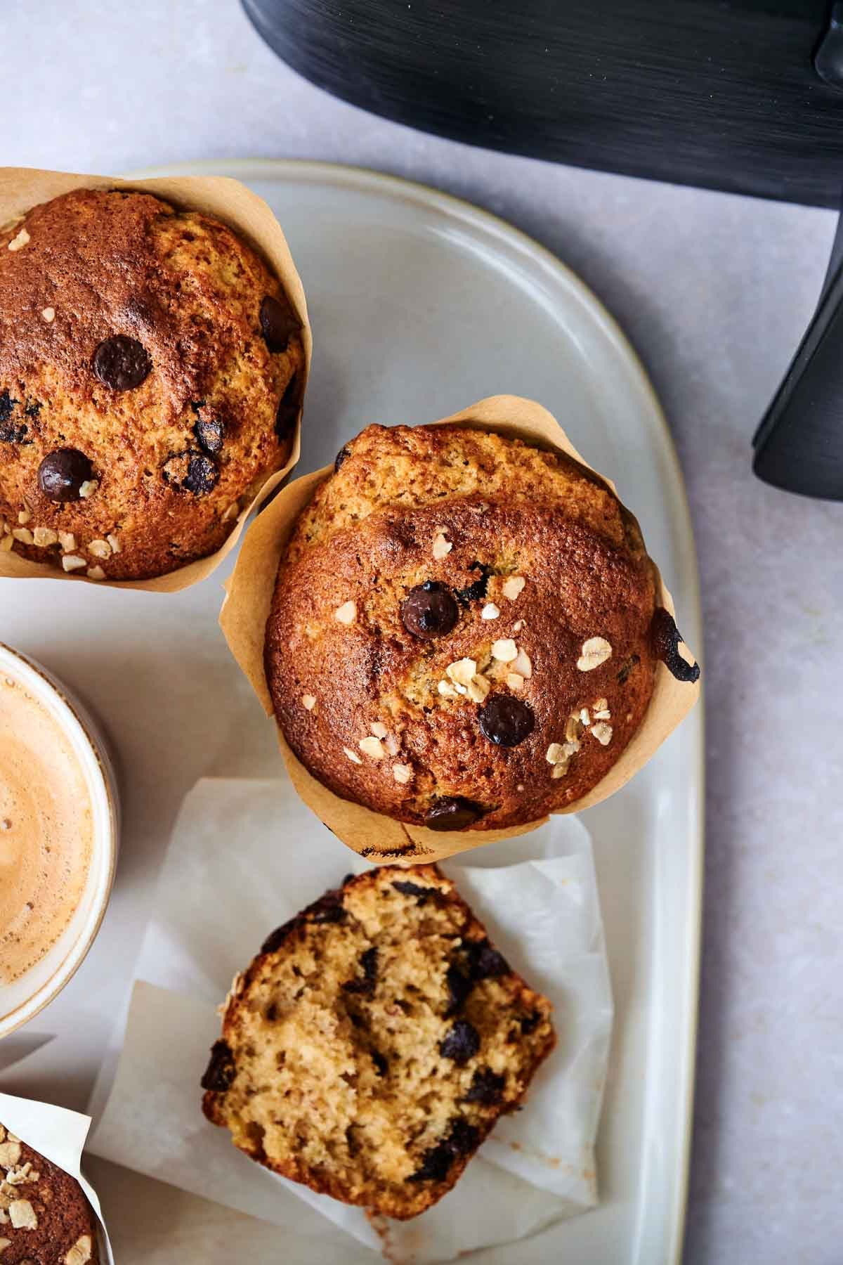 Two Air Fryer Banana Chocolate Chip Muffins and a sliced muffin rest on a white plate, next to a cup of coffee and part of a black appliance.