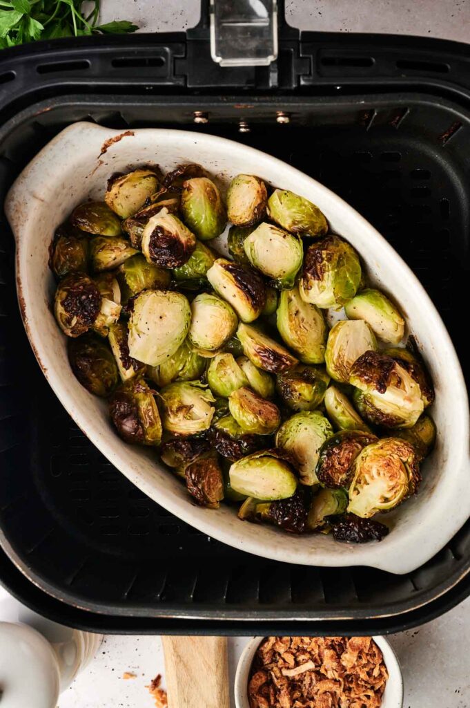A white dish filled with Air Fryer Brussels Sprouts Casserole sits inside an air fryer basket, with crispy bits visible on some sprouts.
