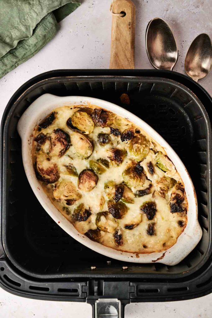 An oval dish of Air Fryer Brussels Sprouts Casserole with melted cheese sits inside an air fryer basket, next to two metal spoons and a green cloth.